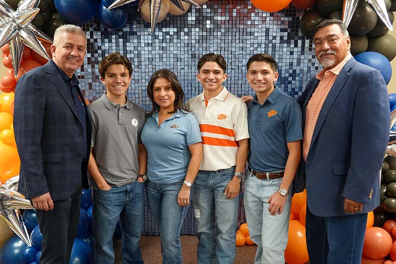 Six attendees pose together in front of a decorative backdrop featuring orange, blue, black, and silver balloons during a UTEP Engineering homecoming event, standing side by side for a group photo while wearing College of Engineering apparel and event name badges.